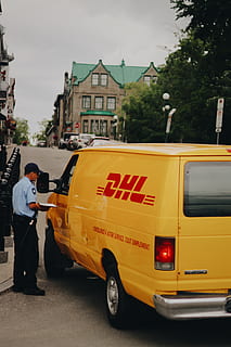 yellow dhl van, van, man, male, dhl, company, logo, postal, postage, mail, shipping, travel, transport, building, architecture, tree, street, road, real people, mode of transportation, transportation, one person, city, full length, built structure, standing, building exterior, men, land vehicle, occupation, outdoors, uniform, day, car, motor vehicle, government, 5K, CC0, public domain, royalty free