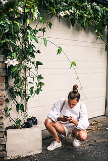 uomo, ducking, bianca, porta del garage, detenzione, smartphone, telecamera, cellulare, luce del giorno, ambiente, flora, fiori, giardino, crescita, edera, indossare, persone reali, pianta, una persona, stili di vita, attività di svago, lunghezza intera, natura, vista frontale, abbigliamento casual, giovane adulto, giorno, foglia, colore verde, parte vegetale, architettura, struttura costruita, seduta, caratteristica della parete, all'aperto, 4K, CC0, dominio pubblico, privo di copyright