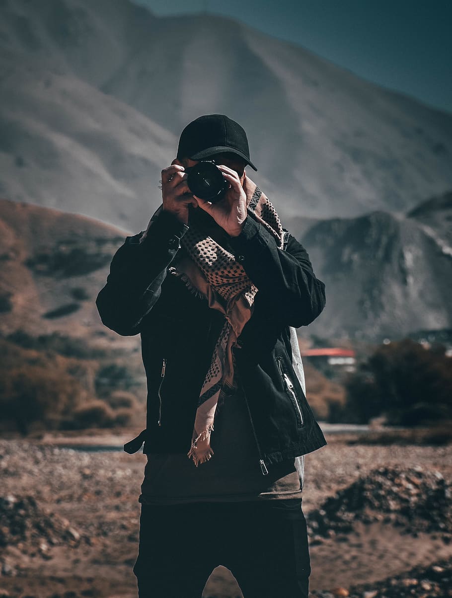 persona che tiene la fotocamera, sfondo sfocato, telecamera, berretto, luce del giorno, paesaggio, uomo, montagne, rocce, sciarpa, suolo, scattare foto, indossare, capi di abbigliamento, una persona, persone reali, tre quarti, vista frontale, concentrarsi sulle conoscenze acquisite, stili di vita, in piedi, attività di svago, natura, escursioni a piedi, giovane adulto, cappello, metà adulto, giacca, avventura, giorno, uomini, vestiti caldi, all'aperto, 4K