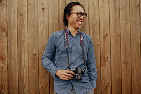 smiling, man, holding, camera, human, person, photography, photographer, clothing, apparel, electronics, photo, pants, strap, glasses, accessory, accessories, shirt, sitting, suspenders, sleeve, jeans, eyeglasses, happiness, one person, young adult, emotion, front view, waist up, cheerful, standing, adult, looking at camera, copy space, fashion, portrait, casual clothing, wood - material, hairstyle, 5K, CC0, public domain, royalty free